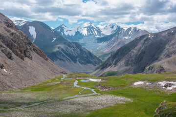 Mountain creek and lake in sunlit green alpine valley against few big snowy pointy peaks far away under clouds in blue sky. Awesome scenic aerial view to three large beautiful snow-capped peaked tops.