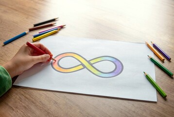 A hand coloring a rainbow infinity symbol on a white sheet of paper with colored pencils scattered...