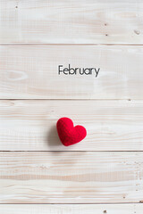 A single red heart placed on the right side of an empty white wooden table, with February in an elegant script font, with space for text or graphics above it