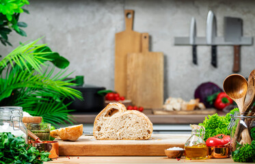 Cut whole grain wheat sourdough bread on wooden cutting board.. Kitchen table with vegetables and herbs against the background of modern classic kitchen