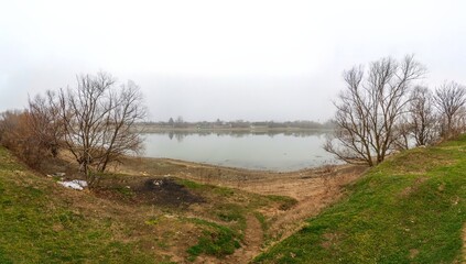 winter bank of the shallow Kuban river with a black fire pit made of old bricks and household garbage - a large panorama on a cloudy morning
