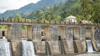 A concrete dam with cascading water in a lush mountainous landscape, featuring a house and trees in the background under a cloudy sky.