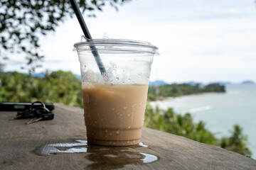 A plastic cup of iced coffee with condensation, placed on a wooden surface overlooking a scenic coastal view with lush greenery and calm ocean waves in the background.