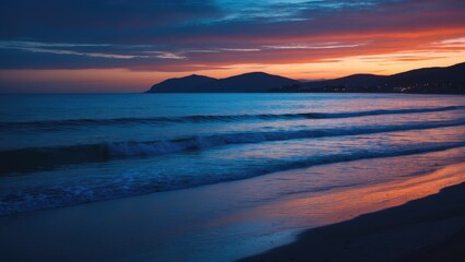 Fototapeta premium Serene coastal sunset with deep blue and orange hues reflecting over calm ocean waves and silhouetted hills on the horizon.