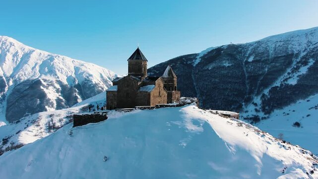 Tourists walking near iconic gergeti trinity church, nestled against snow-capped caucasus mountains in winter, showcasing georgian landscape under bright sunlight