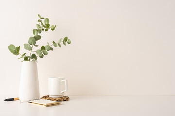 Minimalistic arrangement with a white vase of eucalyptus leaves, coffee mug, and notepad on a clean surface. Highlights cozy, natural, and elegant decor elements for creative and simple living spaces