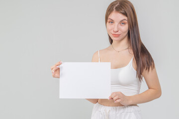 Fototapeta premium Close-up portrait of young brunette with empty A4 sheet of paper on a white background. Copyspace
