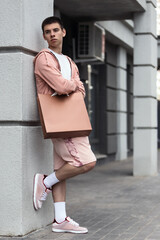 young man poses in trendy summer hoodie in shorts and sneakers with bag outside on sunny warm day in summer, concept of trendy urban summer clothing, youth trend