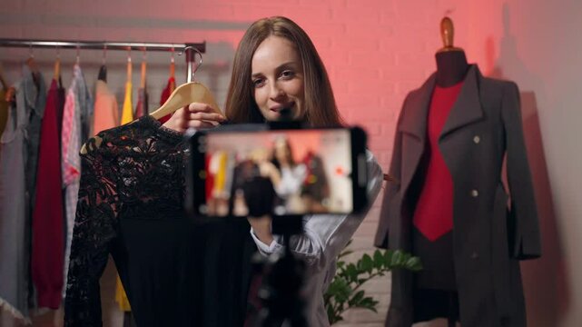 Young pretty female influencer live streaming in her local store using smartphone, businesswoman communicating with virtual audience advertising high quality fashion clothes, blogger recording video