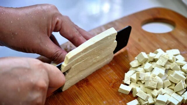 Video footage shows a chef cutting tofu into small pieces on a wooden cutting board to prepare Pad Thai in a home kitchen.