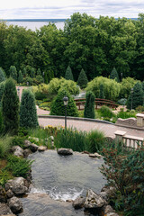 Water flows down an artificial waterfall, creating a pond in a beautifully landscaped park with a variety of trees, shrubs, and decorative elements, offering a tranquil and scenic view