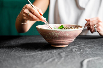 A close-up of a person using chopsticks to enjoy a bowl of colorful noodles topped with fresh herbs, set on a dark table. The scene captures the essence of Asian cuisine.