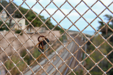Rusted Fence with Locks, Railway Tracks, Mountains, Sea, and Sky in the Background