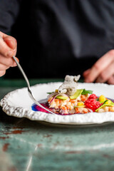 A beautifully plated gourmet dish featuring colorful ingredients, including fresh herbs and sauce, presented on an elegant white plate, with a hand holding a fork, ready to enjoy.