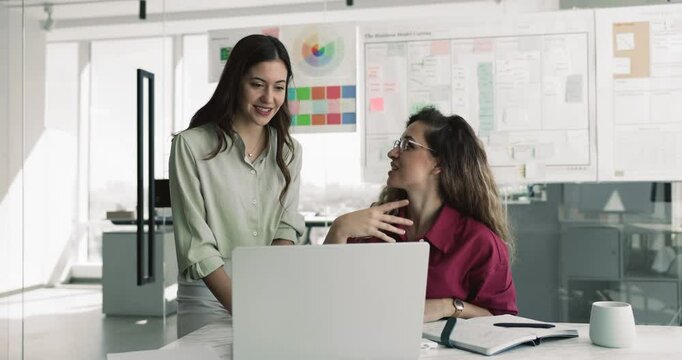 European women workmates talking in office look at laptop screen, review new AI program, work on project, develop strategy feel satisfied and contented with successful, productive workflow, portrait