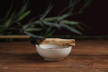 Burnt palo santo stick and bowl on wooden table against black background, closeup