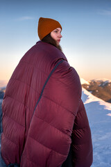 Smiling young woman standing in the background of a winter mountain, wrapped in a warm sleeping bag.