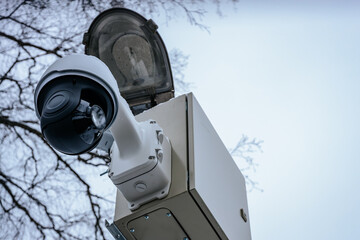 A close-up of a security camera covered in snow, mounted on a metal pole with a control box. Bare...