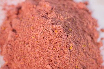 A close-up of a mound of red powdered spices with visible granules and spice particles on a white plate. The variations in texture and color highlight its rich composition.