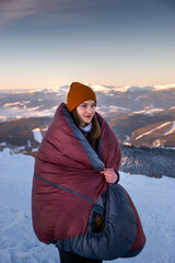 Smiling young woman standing in the background of winter mountains, wrapped in a warm sleeping bag.