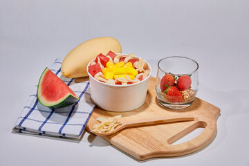 A tropical fruit bowl filled with fresh mango, watermelon, and coconut flakes, served in a white cup on a wooden board. Surrounded by whole mango, watermelon slice, strawberries, and granola.