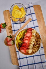 A tropical fruit bowl filled with fresh mango, strawberry, and coconut flakes, served in a white cup on a wooden board. Surrounded by whole mango, watermelon slice, strawberries, and granola.
