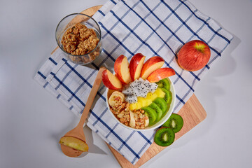 A delicious and nutritious granola bowl topped with fresh apple, kiwi, banana slices, coconut flakes, and almonds. Placed on a wooden board with a checkered napkin.