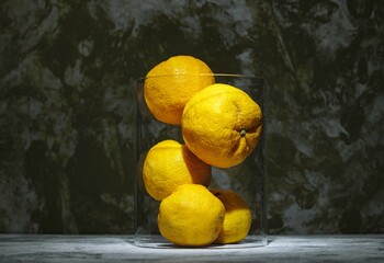 Bright yuzu lemons are carefully arranged within a transparent vessel, showcasing their vibrant yellow hue against an abstract, muted backdrop that highlights their beauty