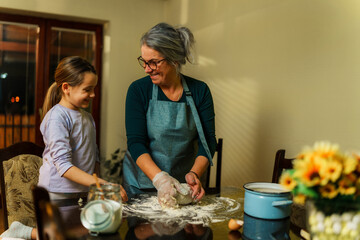 Grandmother, wearing an apron and plastic gloves, kneads dough as her granddaughter watches...