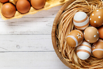 A rustic basket with painted brown and white eggs sits on a wooden surface, with a carton of raw...