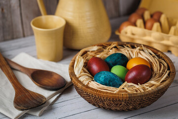 A basket with dyed eggs sits on a rustic table with wooden spoons, ceramic mugs, and fresh bread,...