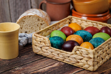 A close-up of a woven basket filled with dyed eggs, surrounded by tableware and bread, creating a cozy, rustic kitchen setting.