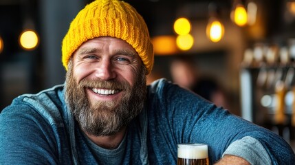 A joyful man with a beard wearing a bright yellow beanie smiles broadly while enjoying a beer in a lively bar setting filled with warm lights and ambiance.