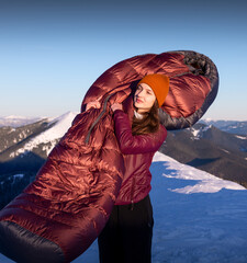 Young woman standing in the background of winter mountains, holding a warm sleeping bag in her...