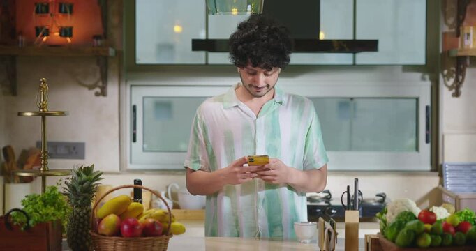 Indian young adult gen z man guy use mobile look chat online app read new text SMS email indoor teen boy hold phone watch funny video enjoy spend day time have fun joy pick up cup drink tea coffee