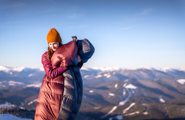 Young woman standing in the background of winter mountains, holding a warm sleeping bag in her...