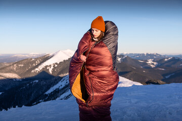 Young woman standing up In a warm sleeping bag without a tent at snowy mountain range at the first...