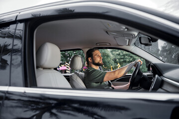 Man driving a car with a thoughtful expression, surrounded by lush greenery and a vibrant outdoor...