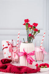 Red carnation and rose flowers in a pink ceramic vase on a light background, next to a mousse cake, a milkshake with red berries and red straws, a heart-shaped cake, Valentine's Day