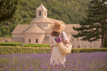 Eine Frau in einem wei&szlig;en Kleid und Hut steht mit dem R&uuml;cken zur Kamera in einem kleinen Lavendelfeld, mit einem wei&szlig;en Haus im Hintergrund.