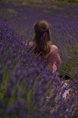 Eine Frau mit offenen Haaren und einem lila Band sitzt in einem Lavendelfeld, mit dem R&uuml;cken zur Kamera. Die Szene ist ruhig und vertr&auml;umt.