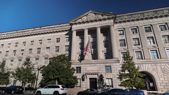 US Department of Commerce building with columns, intricate carvings, and an American flag flying in front. The building has multiple windows, an arched entrance, and is framed by trees.