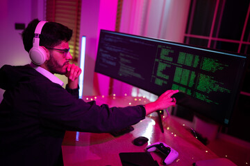 Young man coding in dimly lit room with large screen and gaming console