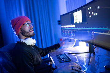 Creative individual editing video content in a dimly lit room using a curved monitor during late...