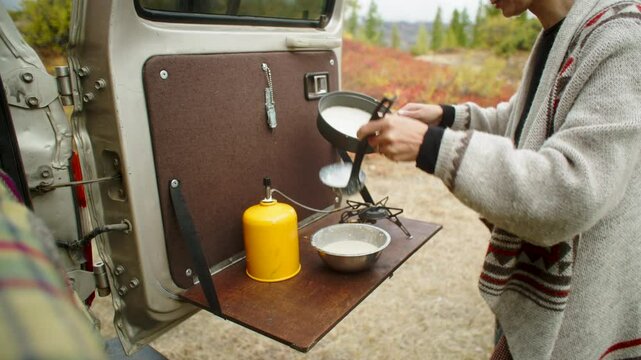 Woman wearing a wool cardigan pouring pancake batter into a pan on a camping stove set up on the tailgate of an off-road vehicle, enjoying a peaceful outdoor cooking experience