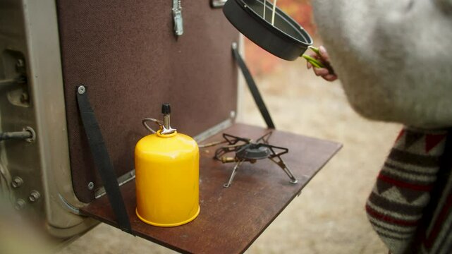 Woman wearing a wool cardigan pouring pancake batter into a pan on a camping stove set up on the tailgate of an off-road vehicle, enjoying a peaceful outdoor cooking experience