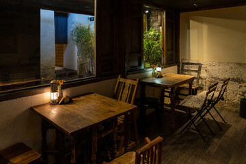 A cozy and atmospheric café at night, featuring rustic wooden tables and chairs illuminated by warm lantern light. The setting exudes a vintage charm with wooden window frames, textured walls, 