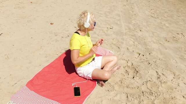 Beautiful cheerful sixty year old blonde woman listening to music in big white headphones sitting on the beach. Woman in yellow T-shirt and white shorts. Pensioner woman in sunglasses