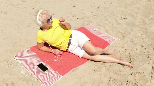 Beautiful cheerful sixty year old blonde woman listening to music in big white headphones sitting on the beach. Woman in yellow T-shirt and white shorts. Pensioner woman in sunglasses