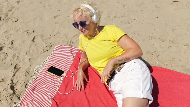 Beautiful cheerful sixty year old blonde woman listening to music in big white headphones sitting on the beach. Woman in yellow T-shirt and white shorts. Pensioner woman in sunglasses